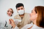 Eine Patientin betrachtet ihr Lächeln im Handspiegel, während ein Zahnarzt mit Mundschutz Zahnfarbmuster danebenhält.