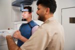 Ein Patient im Röntgengerät, betreut von einem Praxismitarbeiter, der die Einstellungen kontrolliert.