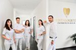 Kamyab Banitorfi und vier Teammitglieder des Dentalogicum Berlin stehen im Praxisflur vor dem Logo der Zahnarztpraxis und posieren gemeinsam für ein Gruppenfoto.