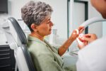 Eine Patientin betrachtet ein Zahnmodell mit Implantat, das ihr vom Zahnarzt im Behandlungsstuhl gezeigt wird.