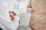 Zahnärztin führt Kontrolluntersuchung bei Patientin durch, Patientin liegt lächelnd auf dem Behandlungsstuhl im hellen Praxisraum