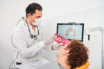 Dr. Kamyab Banitorfi erklärt einem Patienten anhand eines Modells und einer digitalen Darstellung auf dem Bildschirm die Anfertigung einer Zahnschiene