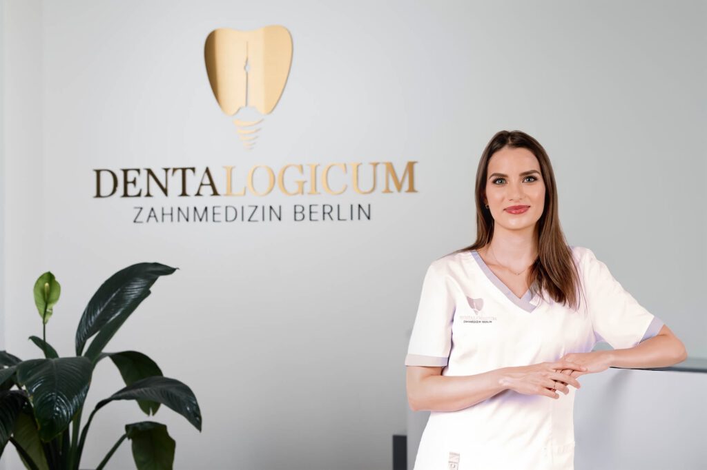 Zahnärztin Corinna Eberle vom Dentalogicum Berlin steht lächelnd im Empfangsbereich der Praxis vor dem goldenen Logo an der Wand.
